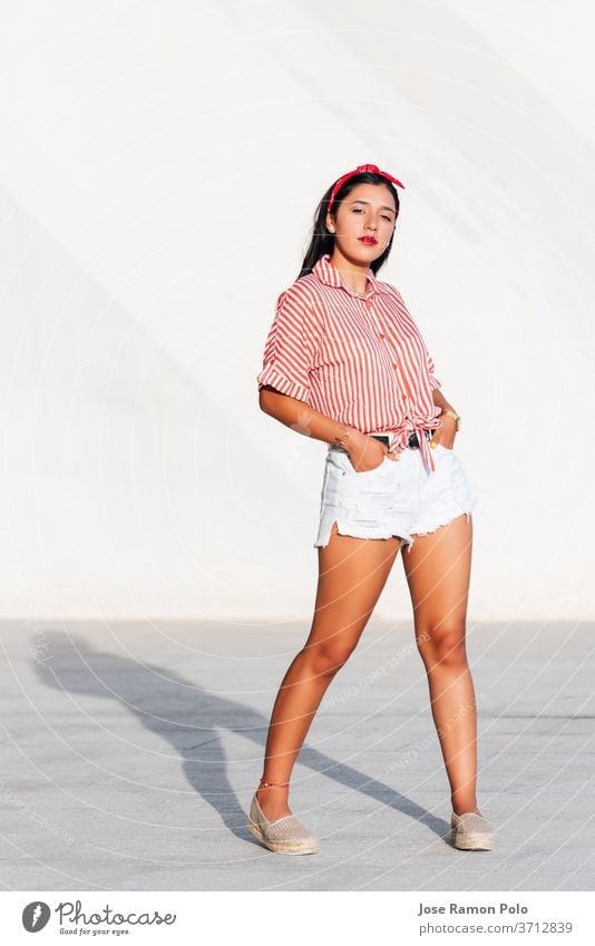 Young Latin girl with black hair and red ribbon in her hair, standing looking at the camera on a sunny day with a white background, wearing a red and white striped shirt and short pants