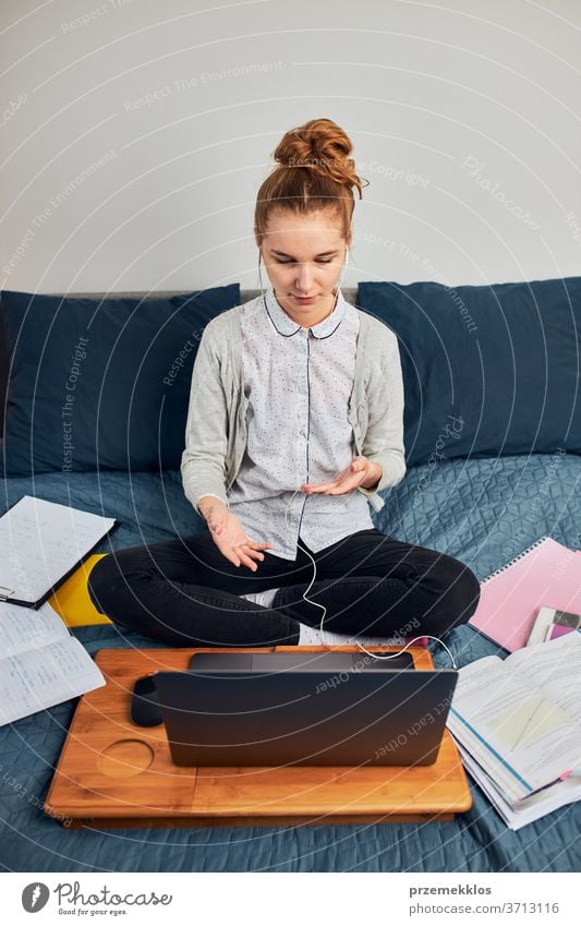 Young woman student having classes, learning online, watching lesson remotely, listening to professor, talking with classmates on video call from home during quarantine. Young girl using laptop, headphones, books, manuals sitting on bed