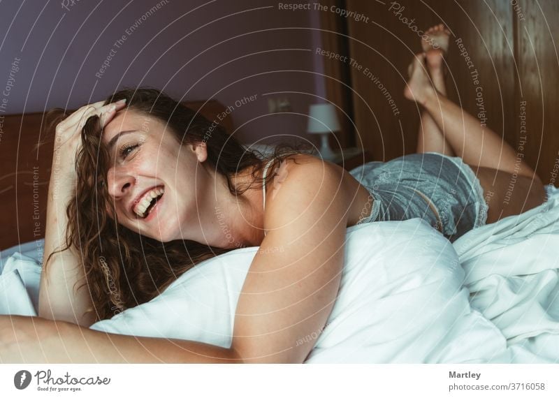 A young woman with brunette hair is lying on her stomach in bed. The girls is laughing and has one arm by her head. Her feet are up in the air. She looks happy.