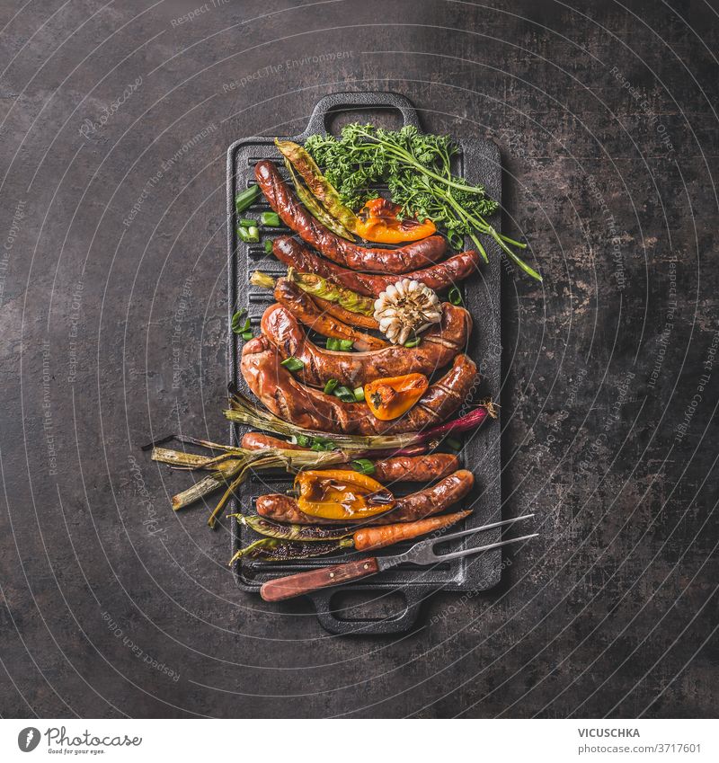 Grilled sausages on cast iron plate. Top view griddle grilled food pepper spring onion garlic paprika herbs carrot meet fork summer barbecue concept dark table