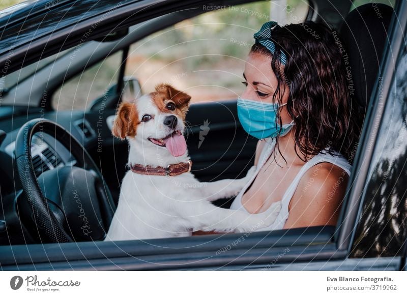 young woman wearing protective mask in a car. cute jack russell dog besides. Travel and new normal concept corona virus travel sun love driveway happy