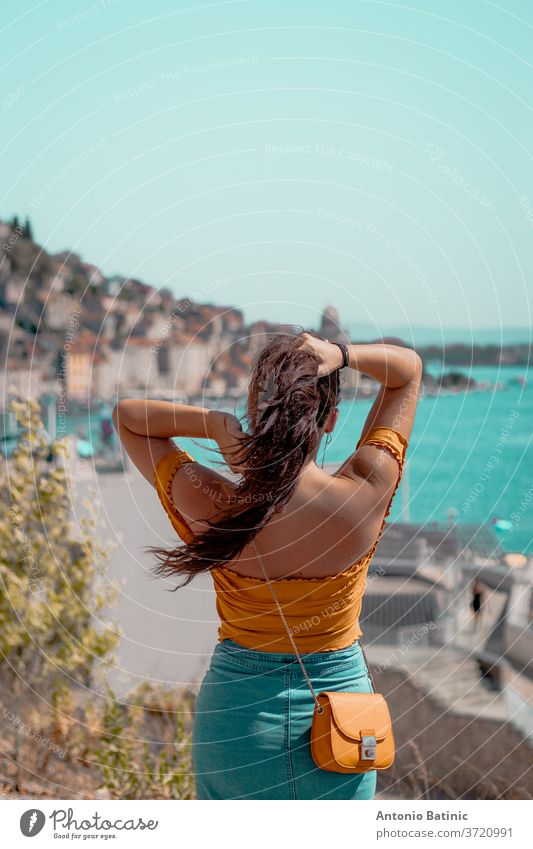 Back view of an attractive brunnete holding her long hair up, looking into the distance. Posing near the bright blue sea, outline of the town of Sibenik in Croatia in the distance
