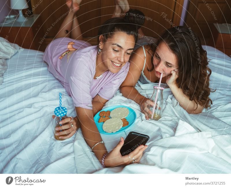 Two girls friends are having breakfast in the bed of hotel room lying together having fun talking and with smartphones browsing internet. woman people indoors