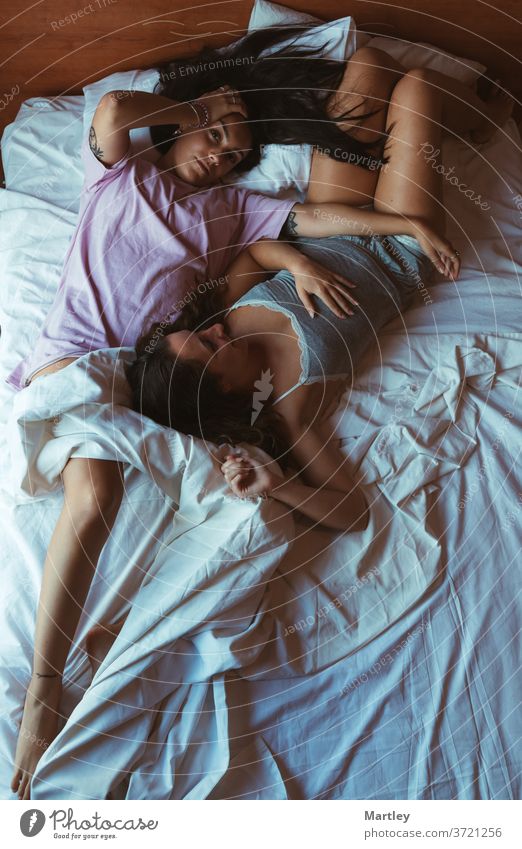 Tender scene of two young women lying in a bed with white sheets cuddling with a thoughtful expression. lesbian selfie at home bedroom indoors love woman pillow