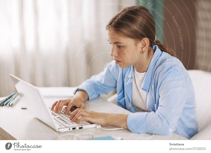 Teenage girl wearing headphones sitting at table and doing homework by video conference using laptop, online education, distance learning, technology concept