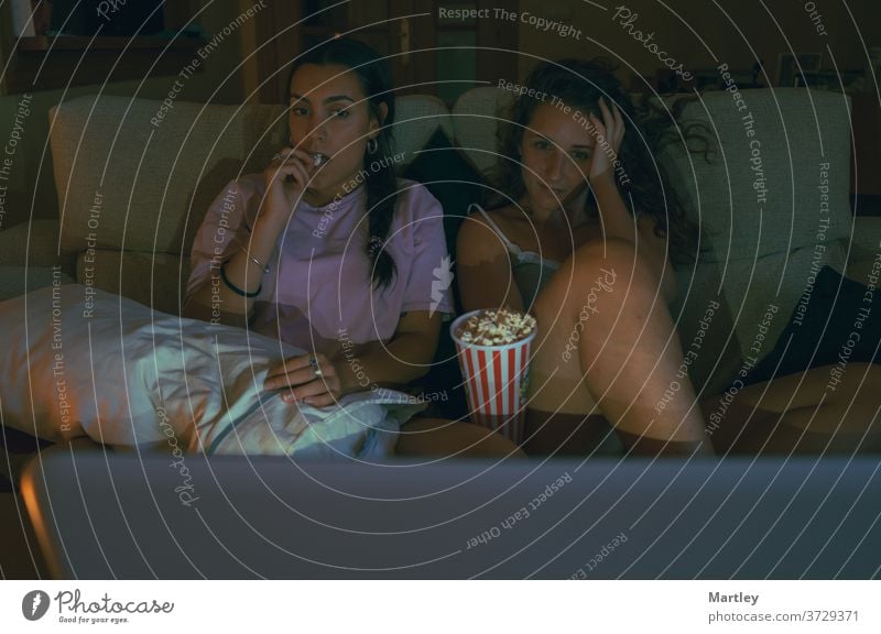 Two teenagers girl eating popcorn and watching movie online while sitting on sofa in cozy living room together. indoors people at home relaxation amusement