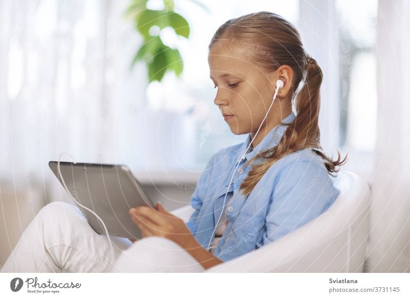 A schoolgirl with headphones sits in a chair with a tablet and does her homework during an online lesson. Online education, Online learning. Social distance, close-up