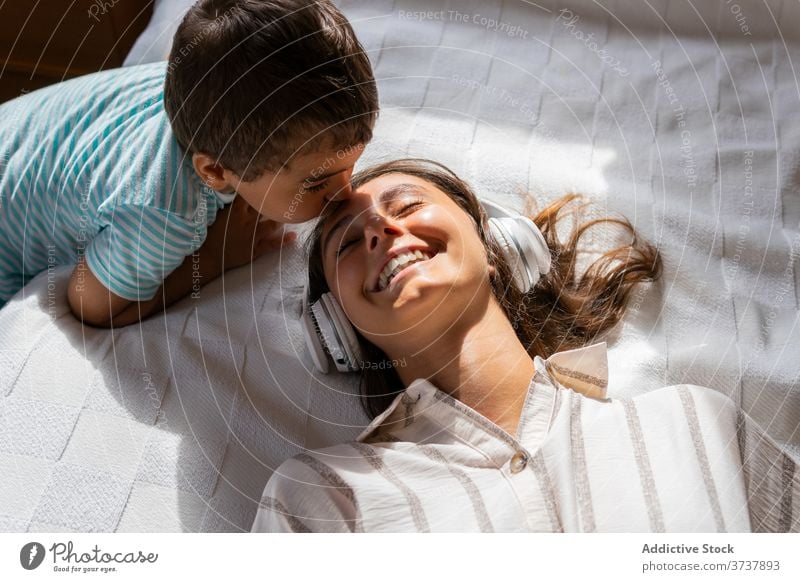 Boy kissing cheerful mother in headphones son listen music happy bed weekend rest love toddler boy child kid woman comfort entertain bedroom lifestyle joy lying