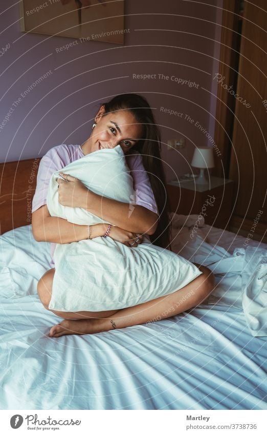 Smiling young woman in sleepwear covering herself with the pillow while sitting and has one arm by her head on comfortable bed and relaxing in morning at home.