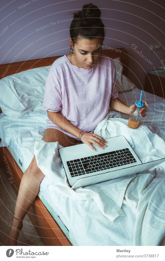 Barefoot young female in pajama teleworking and enjoying refreshing juice while sitting on soft bed and browsing laptop in morning. indoors computer people
