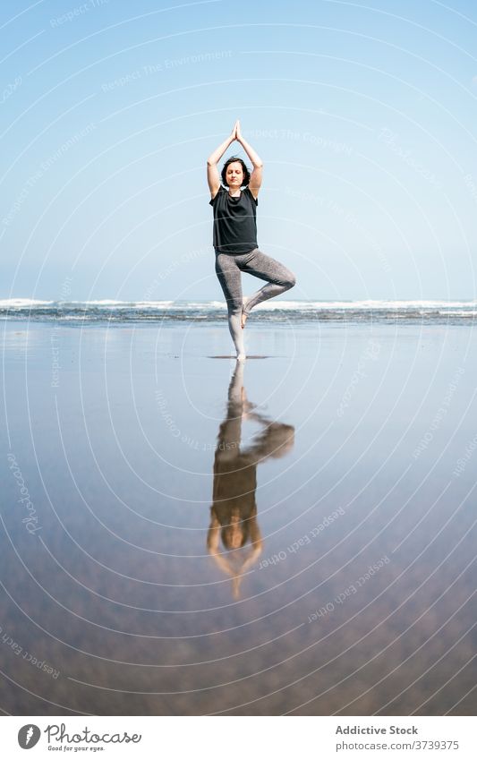Young woman in balancing yoga asana standing on beach sea practice pose tree vrksasana balance calm seashore harmony eyes closed wellness lifestyle female