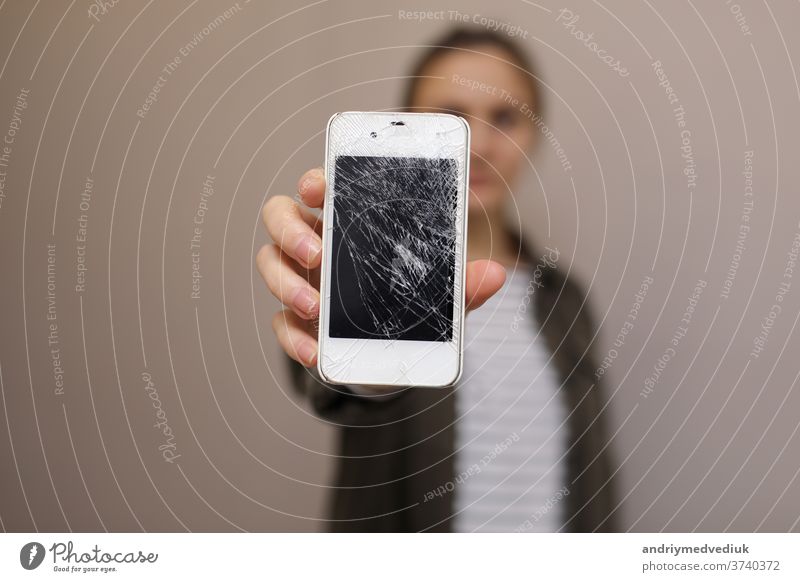 Frustrated young woman holding cellphone with broken screen glass. Phone display needs to repair. Isolated on gray background. selective focus on smartphone