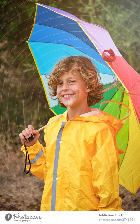 Boy in raincoat holding an umbrella while smiling on camera schoolboy straight pose shower attitude rainbow falling pouring wet weather joy open protection