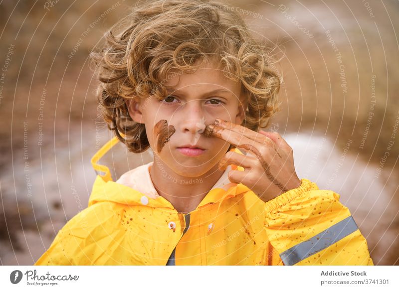 Boy in raincoat drawing marks on his face with the mud facing the camera seriously joke pride infant messy spiritual playful fingers ethnic storm curly makeup
