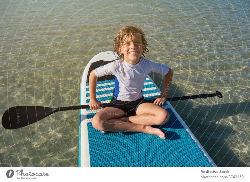 Blonde boy sitting on a paddle surfboard with a paddle floating in the sea lifestyles pose surfing tired comfortable quiet fit recreation joy relaxation model
