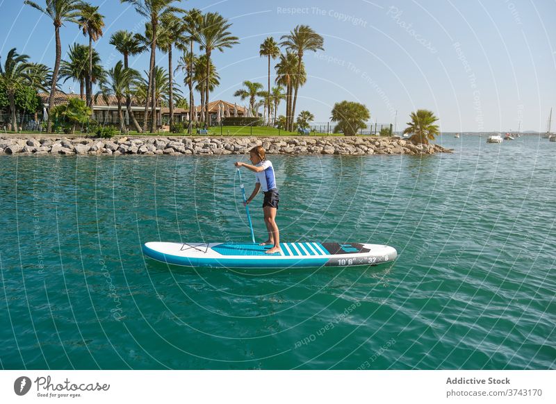 Boy standing on a paddle surfboard rowing in front of a palm beach panorama carefree teenage ride position balance lifestyles enjoyment holidays up wet