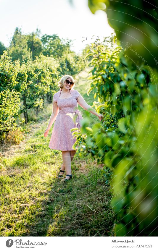 young blonde woman with sunglasses and pink dress walks happily through orchard Young woman Pink Dress Sunset Ease attentiveness closeness to nature pretty