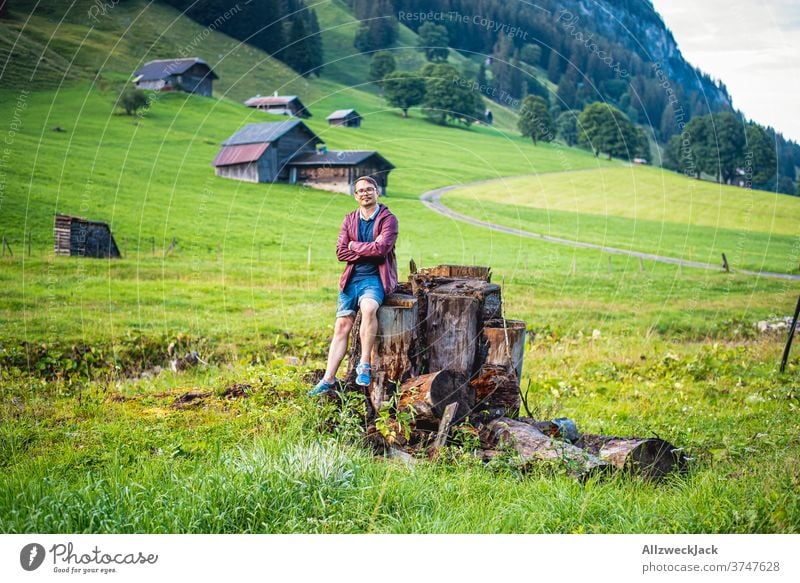 young man with glasses, raincoat and shorts sits on a tree stump on a swiss mountain pasture and looks into the camera Young man Switzerland Alpine pasture