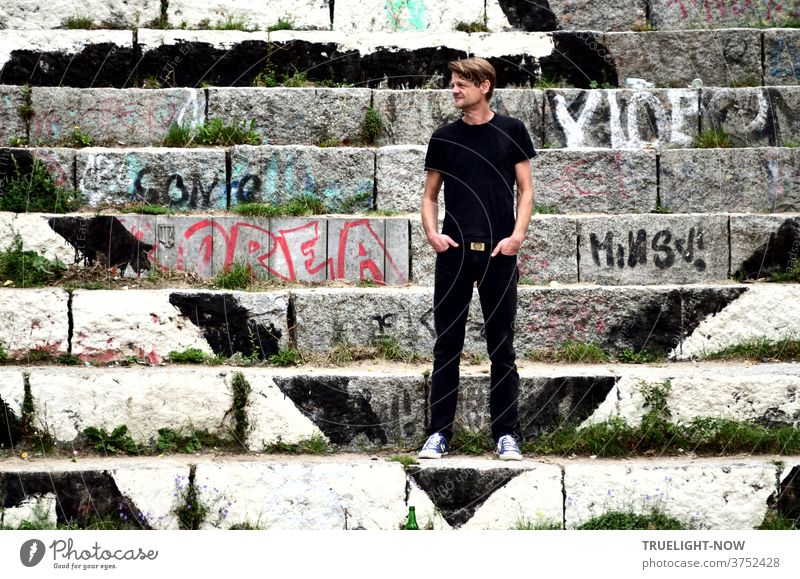 Tim Trzoska, Berlin photographer, Berlin connoisseur by birth, stands in black jeans, black T-shirt and light sneakers on graffiti painted stone steps at Mauerpark in Berlin Prenzlauer Berg