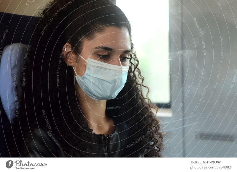 corona thoughts | Young, beautiful up-and-coming model with long, dark, curly hair, dark eyes and fine jewelry on her ear is sitting in the compartment of an ICE train with a protective mask for mouth and nose, looking seriously ahead