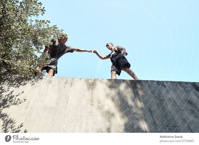 Male friends on high wall in city bump fist greeting men together friendship parkour training prepare stone building cheerful support friendly positive gesture