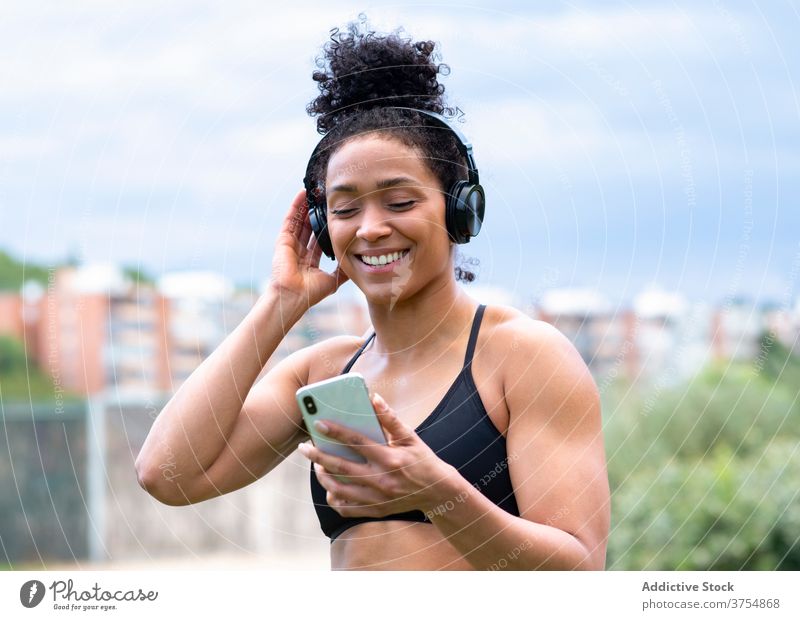 Content sportswoman listening to music in headphones training enjoy athlete fit delight smartphone female ethnic black bra city wireless healthy sportswear