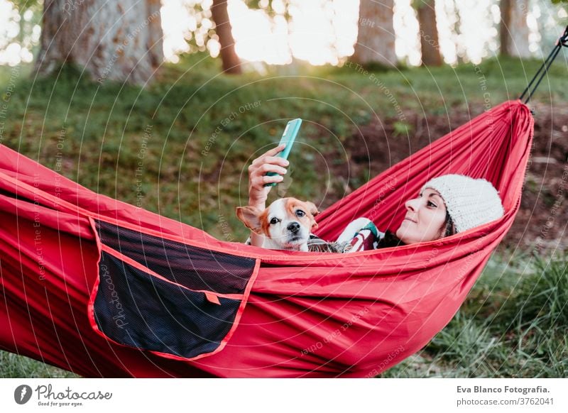young woman relaxing with her dog in orange hammock. Covering with blanket. Using mobile phone. Camping outdoors. autumn season at sunset technology internet