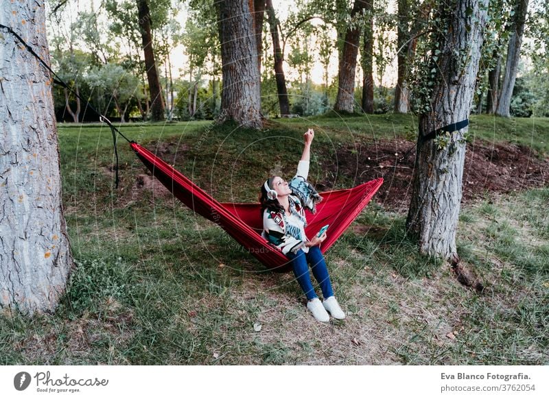 happy woman relaxing in hammock and listening to music on mobile phone and headset. autumn season. camping concept earphones technology internet connection