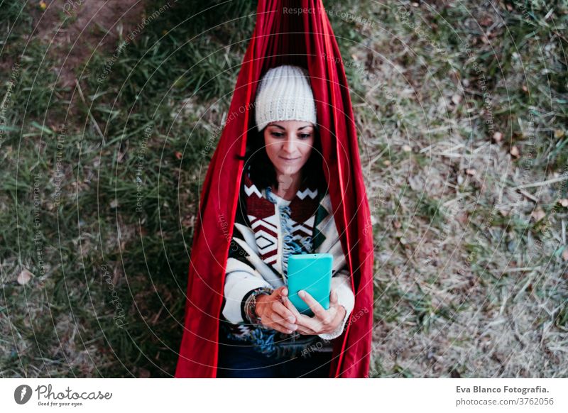 top view of young woman relaxing in hammock using mobile phone. autumn season. camping concept headset earphones listening music technology internet app