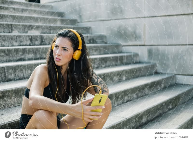 Serious sportswoman listening to music on stairway smartphone headphones rest training workout gadget athlete using mobile device relax urban cellphone step