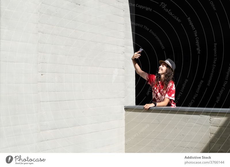 Cheerful young woman taking selfie on smartphone hipster style cheerful urban happy trendy female mobile millennial hat modern building gadget device smile