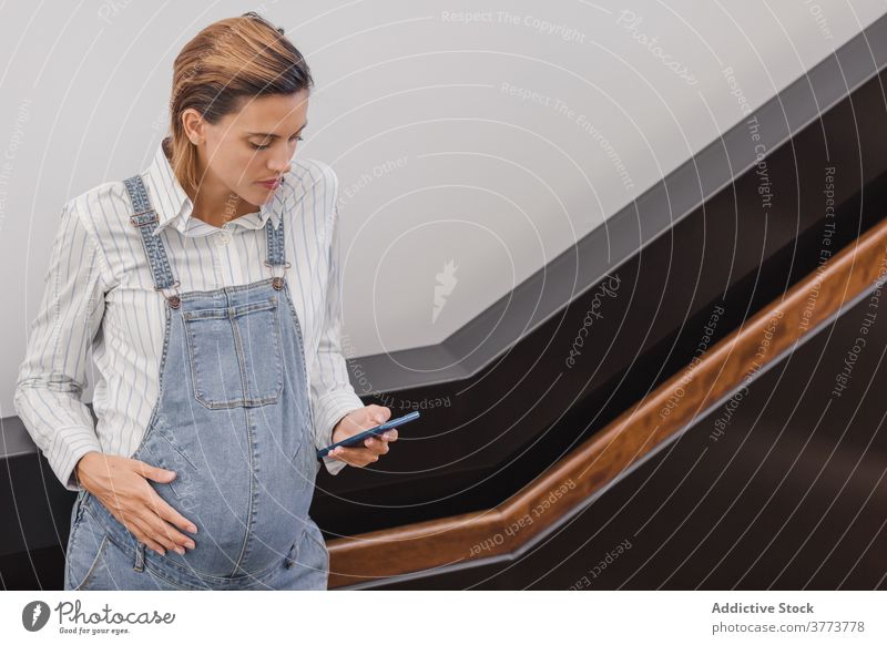 Pregnant woman browsing smartphone in modern building pregnant using message belly maternal pregnancy contemporary female denim staircase device gadget online