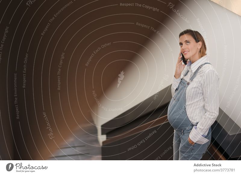 Smiling pregnant woman speaking on cellphone in building smartphone pregnancy tummy modern staircase using female railing conversation lean content gadget