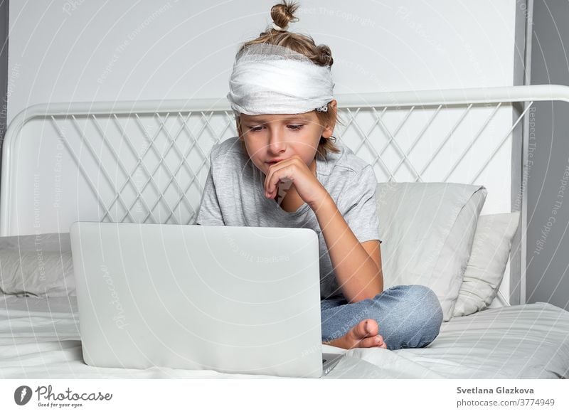Little Caucasian blond boy with a head injury and bandage is sitting on the bed and using laptop. Recovery after incident. Remote communication, distance learning, technologies and gadgets in life