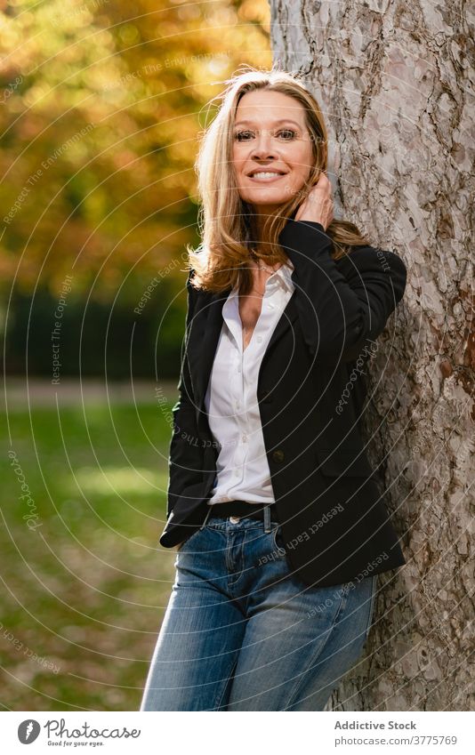 Smiling woman near tree in park stroll relax enjoy season sunlight garden carefree female autumn fall style recreation city casual rest trendy stand lady lean