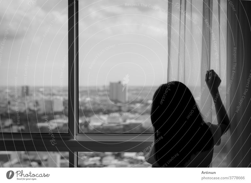 Rear view of woman holding curtain and looking out of glass window. Depressed woman from lock down and quarantine from coronavirus pandemic. Mental health and dementia. Thoughtful woman in dark room.