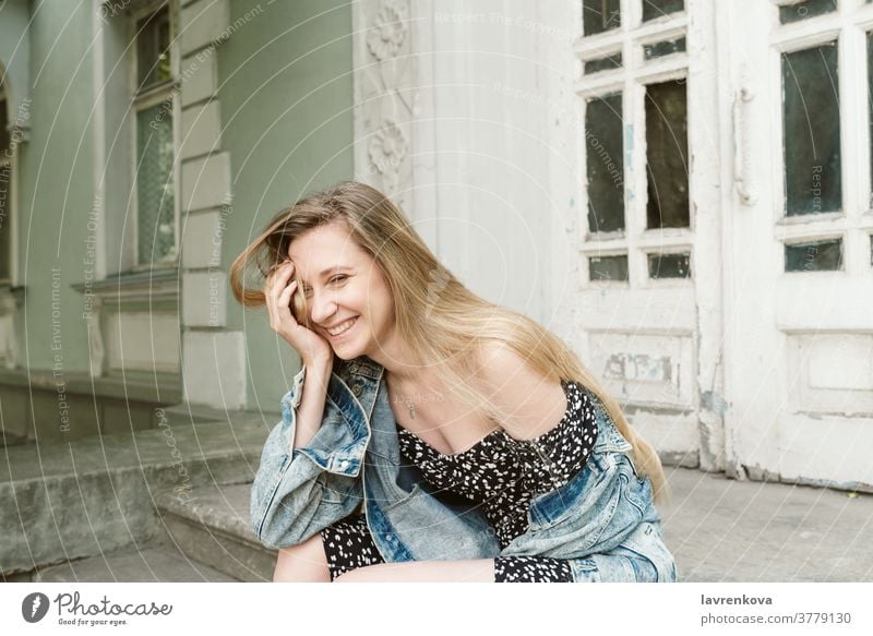 Portrait of laughing blonde female in dress and denim jacket sitting outdoors smiling people lifestyle happy woman girl attractive joyful caucasian adult person