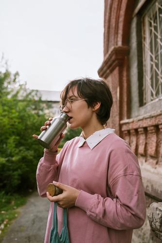 Young adult female with short hair drinking from metal sustainable water outdoors, selective focus bottle hands closeup fingers holding woman girl pink