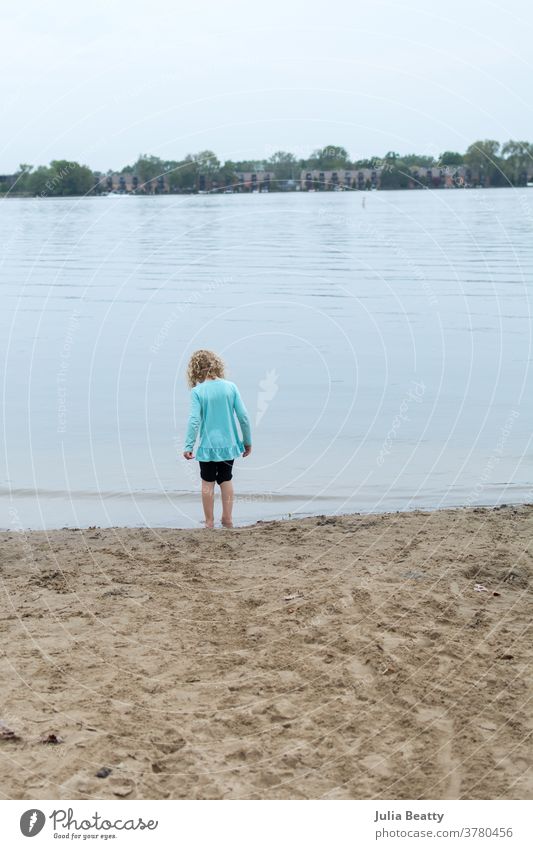 Little girl with curly blond hair alone on lake front beach lakefront sand water ripple child kid Curly hair Blonde blonde hair isolation wading blue soft
