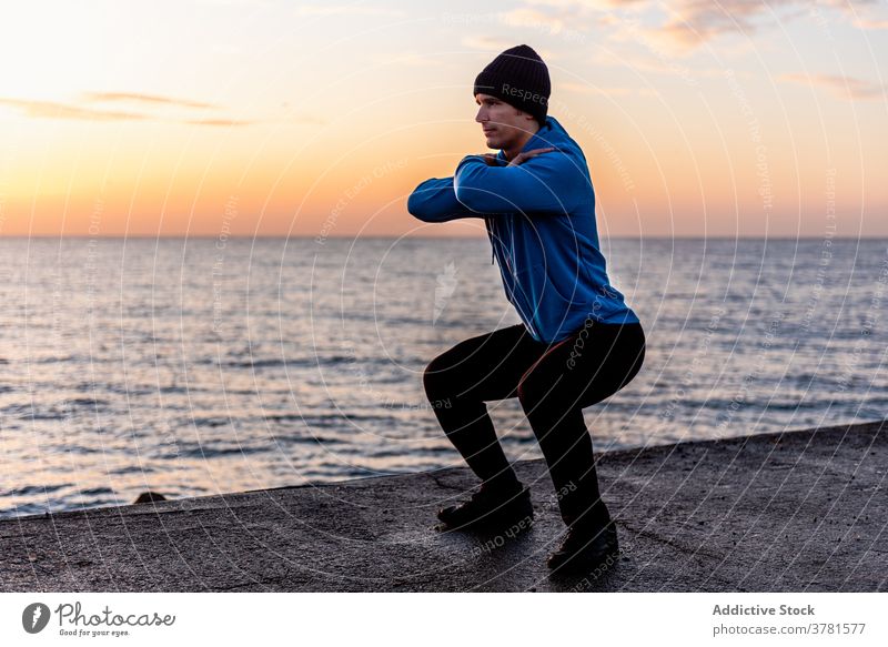Sportsman squatting on city embankment training sea workout fitness exercise sporty urban ocean active male ethnic healthy strong warm up determine motivation