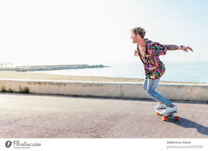 Carefree man riding skateboard along promenade skater summer hipster embankment weekend balance skill male style trendy outfit outstretch young fun happy