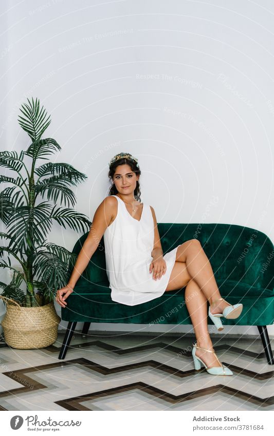Smiling woman in elegant apparel sitting on sofa dress white grace living room relax style delight tender female couch soft trendy summer outfit fashion happy