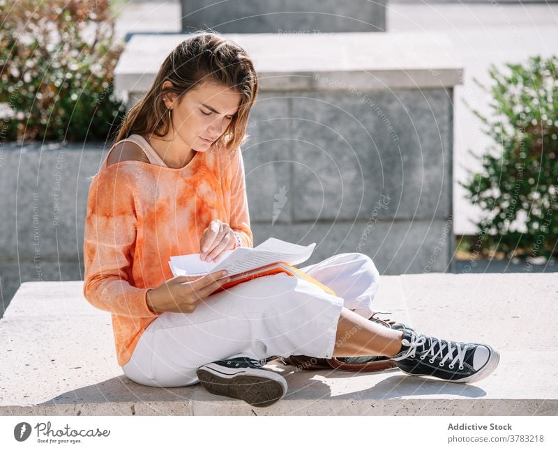 Young student sitting on border opening notebook woman university homework education street study female read legs crossed learn madrid spain young casual