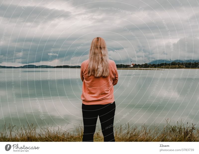 Young woman stands in front of the Forggensee in Füssen Blonde Rear view Relaxation Break Freedom Summer Sun Reflection Lake Lakeside pretty Clouds Sky