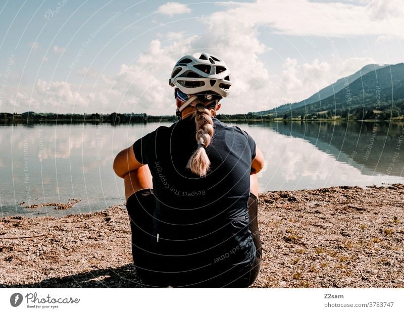 Young cyclist sitting in front of a lake in Allgäu Young woman Blonde Braids Helmet cycling jersey Rear view relaxation Break Relaxation Freedom Warmth Summer
