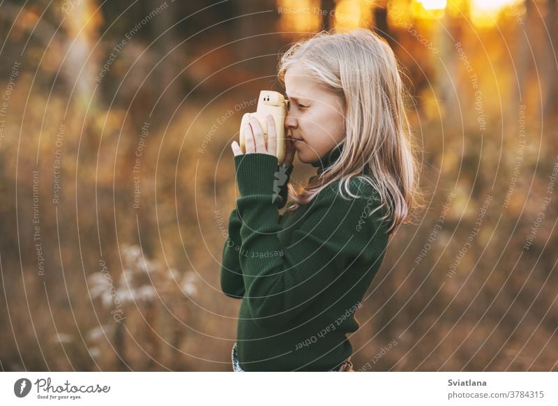 A beautiful teenage girl with blond hair takes pictures of nature with a yellow polaroid camera in an autumn park. Side view. sad pensive child blonde leaf