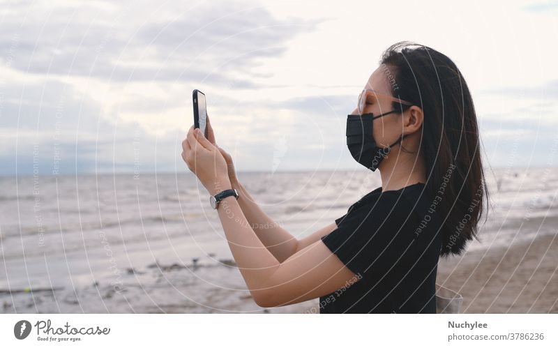 Young asian woman wearing medical mask and taking photos by smartphone at the beach after coronavirus, new normal lifestyle and travel bubble concept attractive