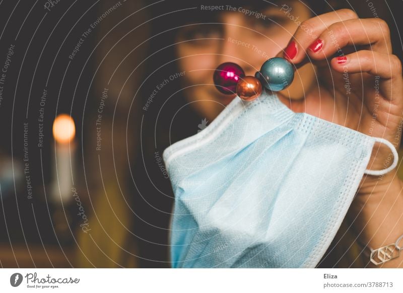 A woman holds a disposable mask decorated with colorful Christmas tree balls. Concept for Christmas during the Corona Pandemic / Corona Thoughts corona Mask