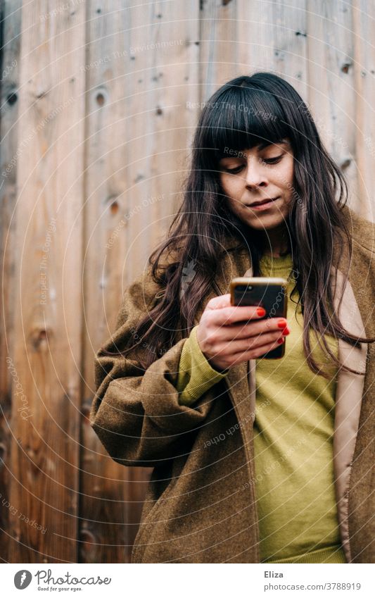 Young woman with long brown hair and autumnal clothes leans against a wooden wall and uses her smartphone Woman out Wooden wall Autumnal Cellphone long hairs