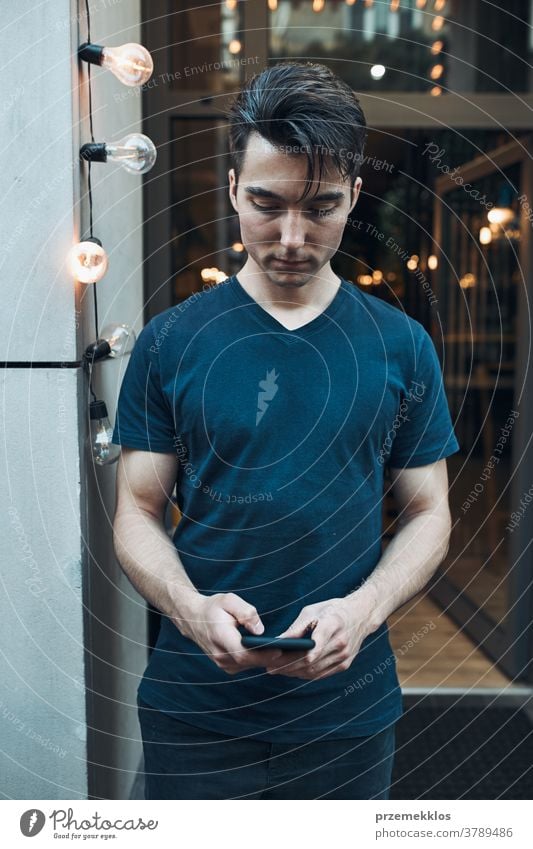 Man standing in front of restaurant in the city center holding smartphone looking at screen boy caucasian downtown evening lifestyle male man outdoors people